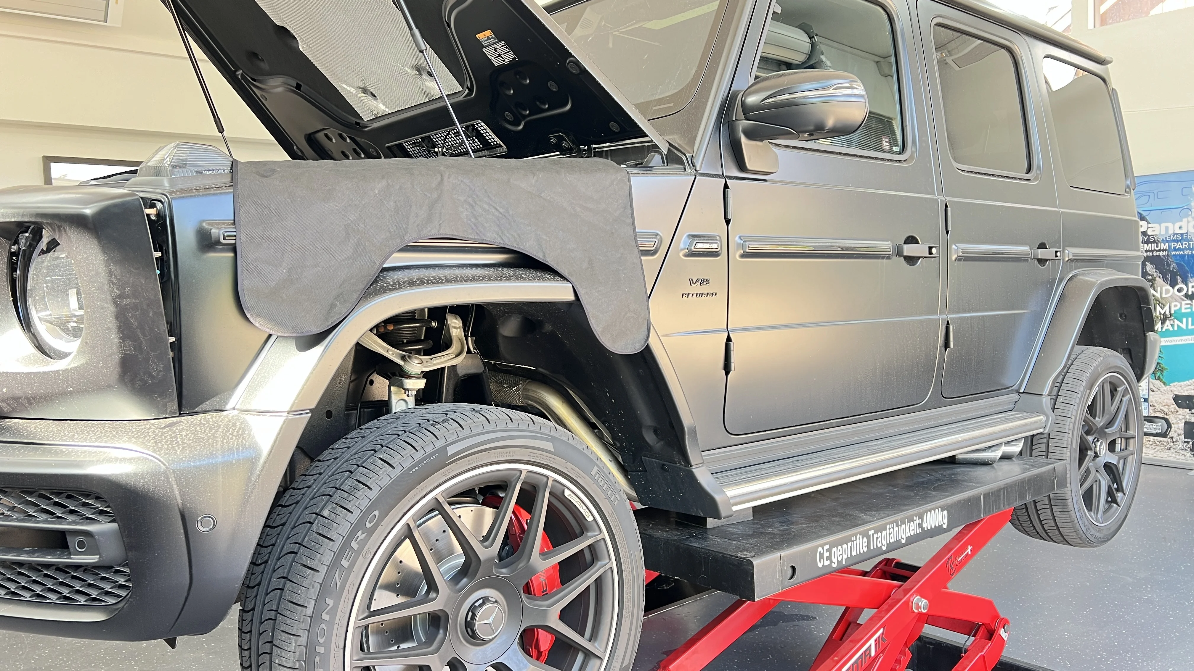 Mercedes G-Klasse auf der Hebebühne mit geöffneter Motorhaube und Schutzmatte über dem Kotflügel