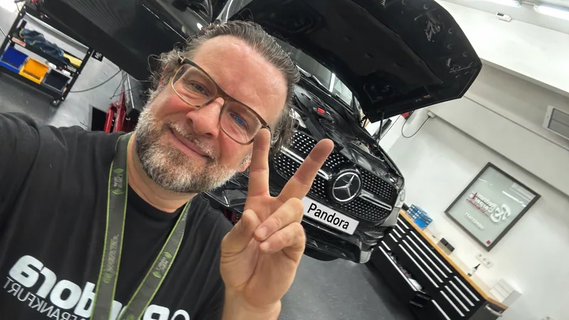 Marco Nolte lächelt in seiner Werkstatt vor einem Fahrzeug und zeigt ein Victory-Zeichen